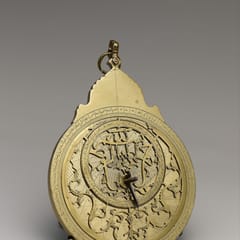 A brass astrolabe with an ornate suspension crown, its pierced star-map plate laced with curling pointers above engraved Arabic script.