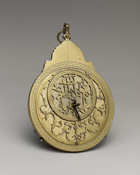 A brass astrolabe with an ornate suspension crown, its pierced star-map plate laced with curling pointers above engraved Arabic script.