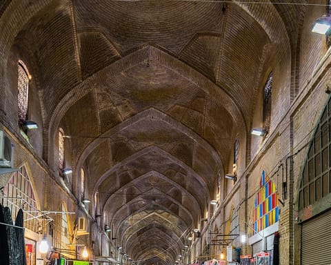 A long vaulted brick bazaar corridor lined with shops of bright folded fabrics, its arches lit by hanging lamps and a window of colored glass.
