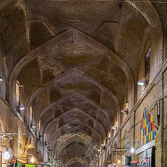 A long vaulted brick bazaar corridor lined with shops of bright folded fabrics, its arches lit by hanging lamps and a window of colored glass.