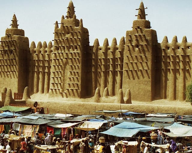 The earthen walls and towers of the Great Mosque of Djenné, bristling with wooden beams, rise above the bright awnings of the market in front of it.