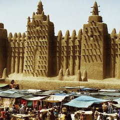 The earthen walls and towers of the Great Mosque of Djenné, bristling with wooden beams, rise above the bright awnings of the market in front of it.