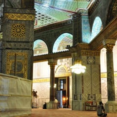 Marble columns with gilded capitals and arches of glittering mosaic surround the ambulatory of the Dome of the Rock, lit by a hanging chandelier.