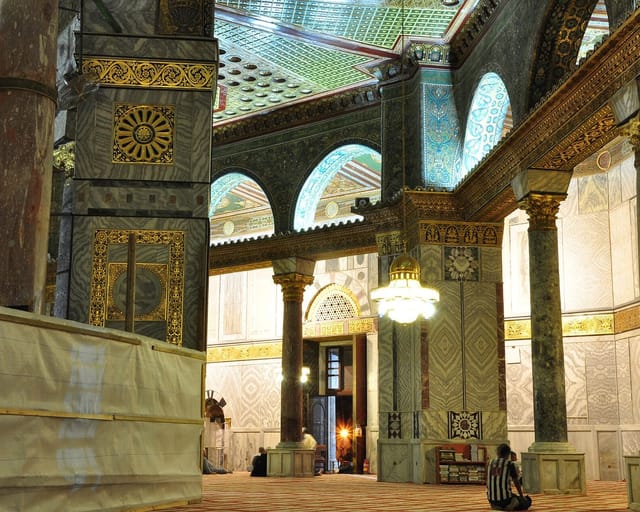 Marble columns with gilded capitals and arches of glittering mosaic surround the ambulatory of the Dome of the Rock, lit by a hanging chandelier.
