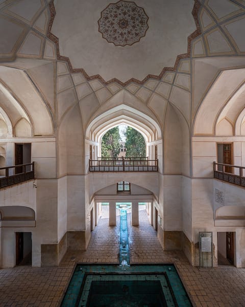 Inside a white garden pavilion with carved vaults and wooden balconies, a turquoise water channel runs out through an arch toward the trees of a Persian garden.