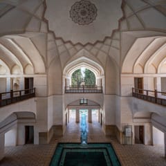 Inside a white garden pavilion with carved vaults and wooden balconies, a turquoise water channel runs out through an arch toward the trees of a Persian garden.