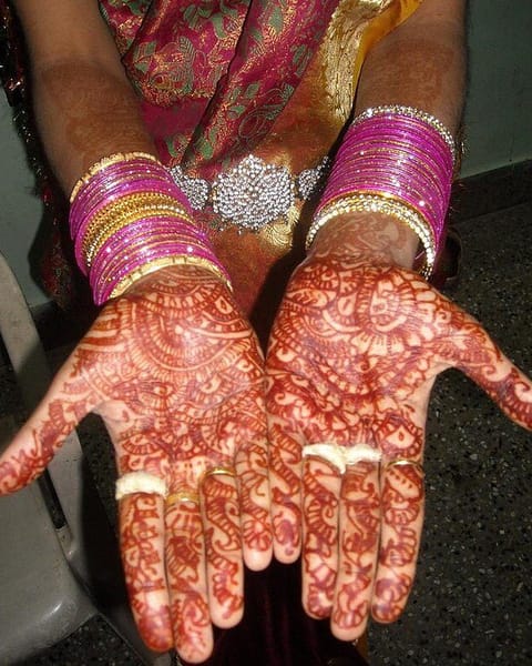 A bride's open palms and fingers covered in fine red-brown henna patterns, with stacks of pink and silver bangles at her wrists.