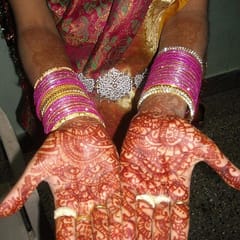 A bride's open palms and fingers covered in fine red-brown henna patterns, with stacks of pink and silver bangles at her wrists.