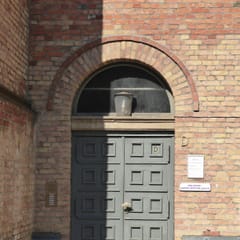 A tall green double door under a round brick arch in a yellow-brick church building, with a small sign for the Ibn Rushd-Goethe Mosque beside it.