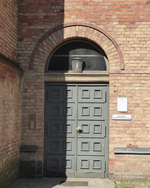 A tall green double door under a round brick arch in a yellow-brick church building, with a small sign for the Ibn Rushd-Goethe Mosque beside it.