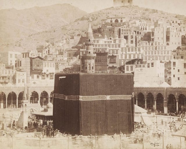 An early sepia photograph of the Kaaba in its black cloth cover inside the arcaded courtyard of the Great Mosque of Mecca, with the houses of the city climbing the hills behind.