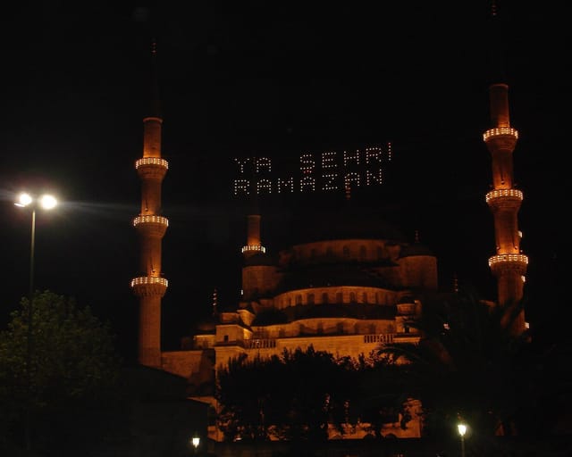 The domes and minarets of the Blue Mosque glow against the night sky, with the words “Ya Şehr-i Ramazan” (O month of Ramadan) spelled in lights between the minarets.