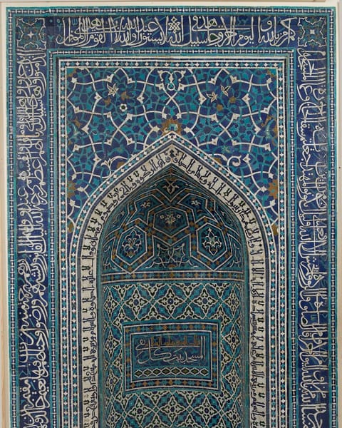 A tall prayer niche in turquoise, cobalt, and white tile mosaic, framed by bands of Quranic inscription with a pointed arch at its heart.