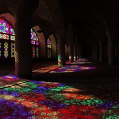 Sunlight through stained-glass windows throws pools of red, blue, green, and violet across the carpets between the carved columns of a dim prayer hall.