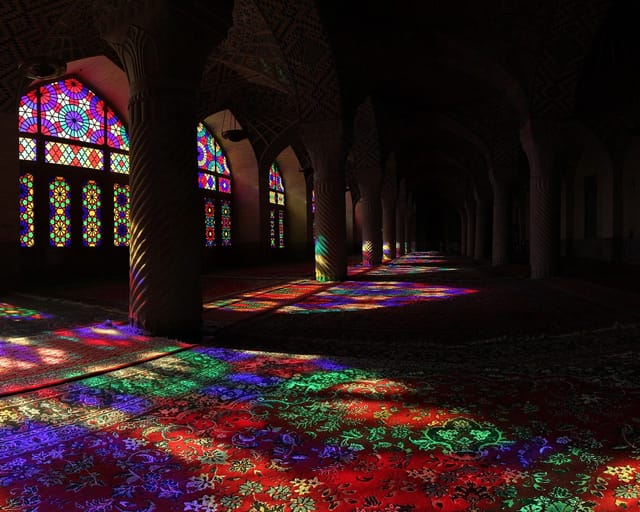 Sunlight through stained-glass windows throws pools of red, blue, green, and violet across the carpets between the carved columns of a dim prayer hall.