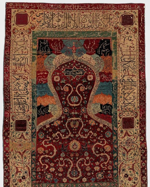 A silk and wool prayer rug with an arched niche filled with red flowering vines, framed by wide borders of woven inscription.