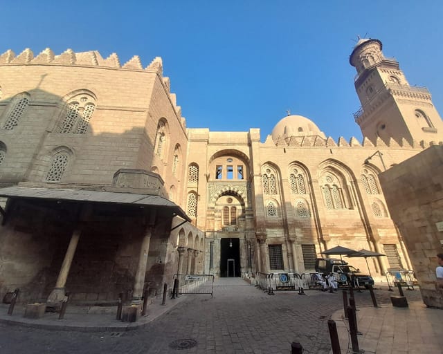 The tall stone facade of a Mamluk hospital, madrasa, and mausoleum complex, with pointed-arch windows, a dome, and an ornate minaret against a blue sky.