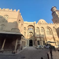 The tall stone facade of a Mamluk hospital, madrasa, and mausoleum complex, with pointed-arch windows, a dome, and an ornate minaret against a blue sky.