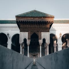 A carved wooden pavilion with a green-tiled roof and stucco arches stands at the end of a long, still water channel in a white arcaded courtyard.