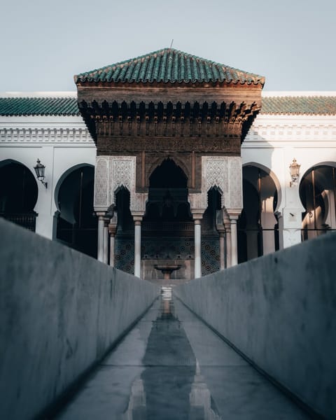 A carved wooden pavilion with a green-tiled roof and stucco arches stands at the end of a long, still water channel in a white arcaded courtyard.