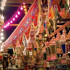 Dozens of colored-glass Ramadan lanterns hang in a market stall at night beside red and blue appliqué tent cloth and a string of pink lights.