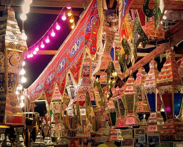 Dozens of colored-glass Ramadan lanterns hang in a market stall at night beside red and blue appliqué tent cloth and a string of pink lights.