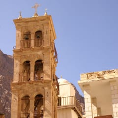 The stone bell tower of a desert monastery rises against a blue sky, and just behind it stands the white minaret of the mosque inside the monastery walls.