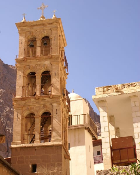 The stone bell tower of a desert monastery rises against a blue sky, and just behind it stands the white minaret of the mosque inside the monastery walls.