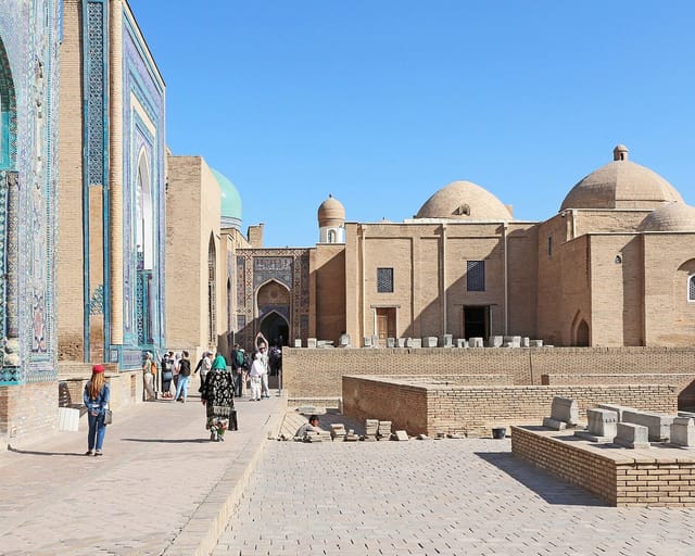 A paved lane of mausoleums in Samarkand: turquoise and cobalt tiled facades on one side and pale brick domed tombs and gravestones on the other.