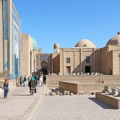 A paved lane of mausoleums in Samarkand: turquoise and cobalt tiled facades on one side and pale brick domed tombs and gravestones on the other.