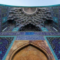 Looking up into the entrance vault of a mosque: a honeycomb of muqarnas tiles in turquoise, cobalt, and gold rises above walls of floral blue tilework.