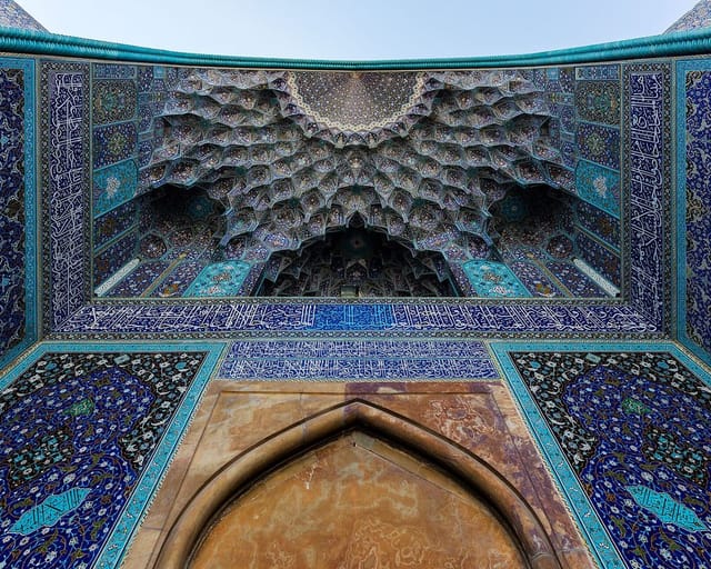 Looking up into the entrance vault of a mosque: a honeycomb of muqarnas tiles in turquoise, cobalt, and gold rises above walls of floral blue tilework.