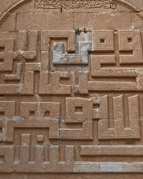 A sandstone wall carved with blocky square Kufic letters, a maze-like grid of right angles framed by a pointed arch.