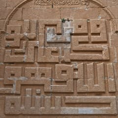A sandstone wall carved with blocky square Kufic letters, a maze-like grid of right angles framed by a pointed arch.