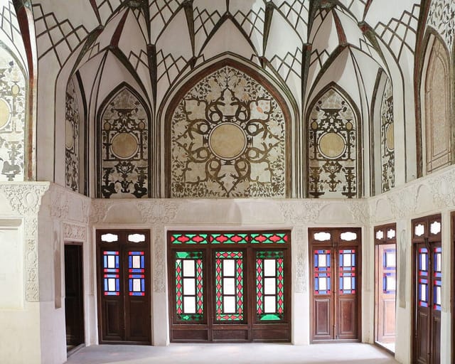 A white reception room with carved stucco vaults and wooden doors set with small panes of red, blue, and green glass.