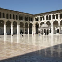A wide marble courtyard shines in the sun, surrounded by two-story arcades, with a small domed ablution fountain near the center.