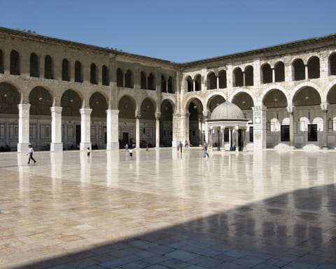 A wide marble courtyard shines in the sun, surrounded by two-story arcades, with a small domed ablution fountain near the center.