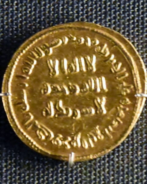 A gold coin on dark cloth with Arabic inscriptions in plain Kufic letters across its face and around its rim.