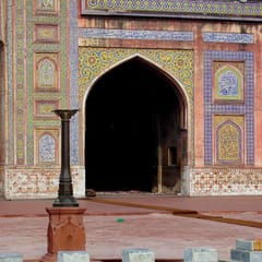 A still ablution pool with marble steps and a slender fountain stand reflects the tiled, pointed-arch entrance of a Mughal mosque.