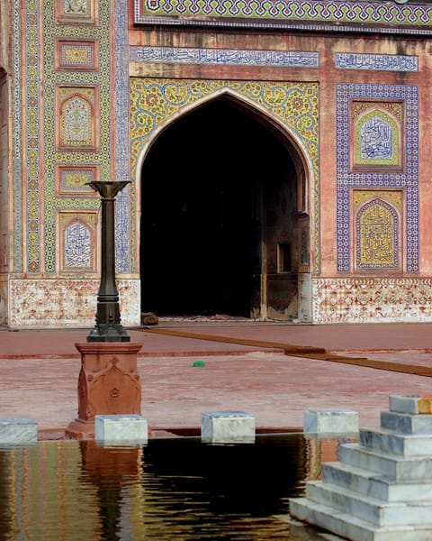 A still ablution pool with marble steps and a slender fountain stand reflects the tiled, pointed-arch entrance of a Mughal mosque.