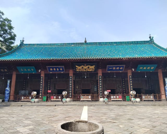 A Chinese-style prayer hall with a sweeping turquoise-tiled roof, red columns, and blue-and-gold signboards under the eaves, fronted by a paved stone courtyard.