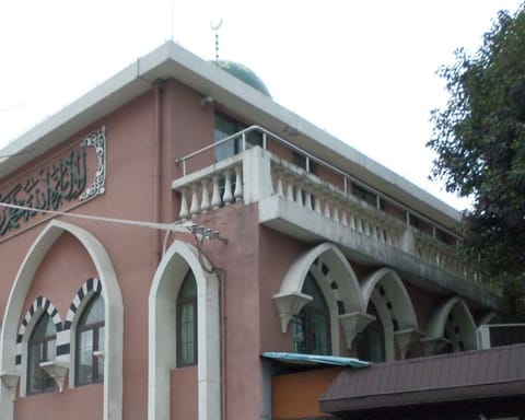 A pink building with tall white pointed arches, a balustraded upper floor, and an Arabic inscription panel under its roofline, beside a leafy tree.