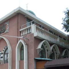 A pink building with tall white pointed arches, a balustraded upper floor, and an Arabic inscription panel under its roofline, beside a leafy tree.